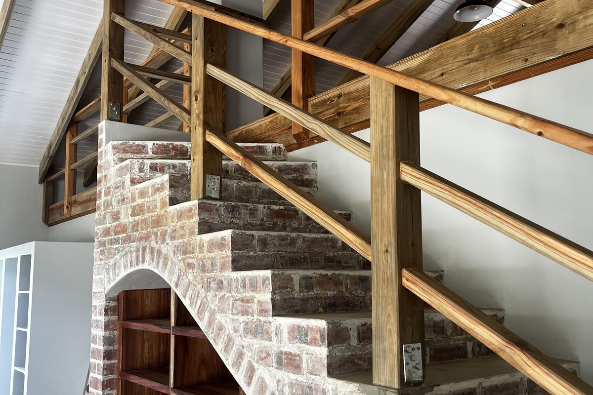 A spacious interior area featuring a unique brick staircase with integrated timber shelving, exposed roof trusses, and high vaulted ceilings.