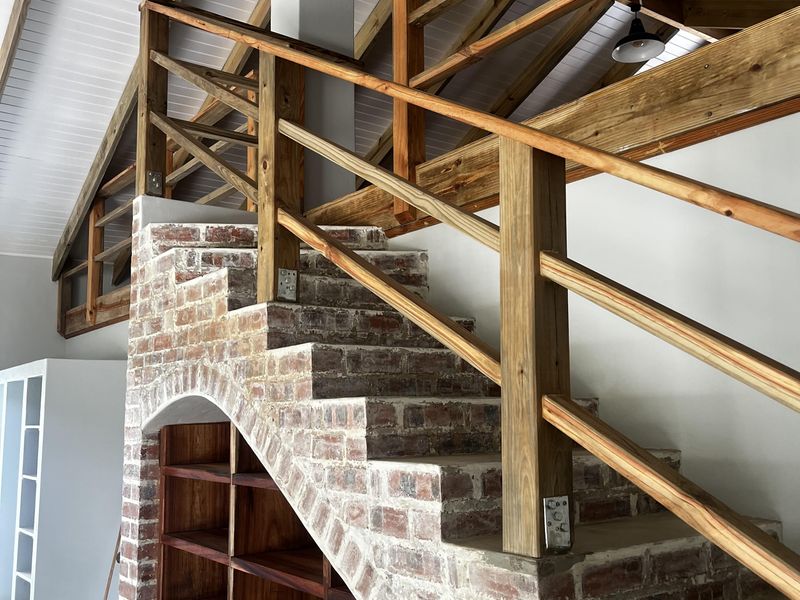 A spacious interior area featuring a unique brick staircase with integrated timber shelving, exposed roof trusses, and high vaulted ceilings.