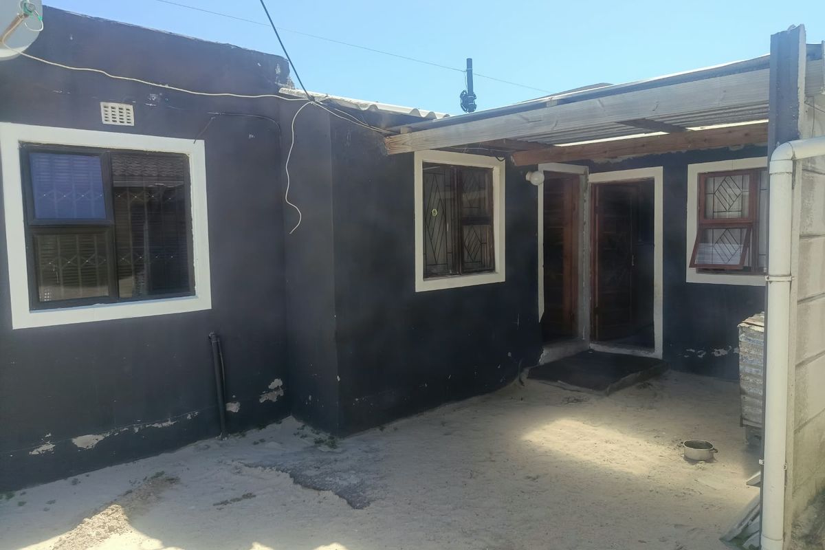 Rear exterior view of the property featuring a covered patio area, dark painted walls with security-fitted windows, and an unpaved sandy courtyard.