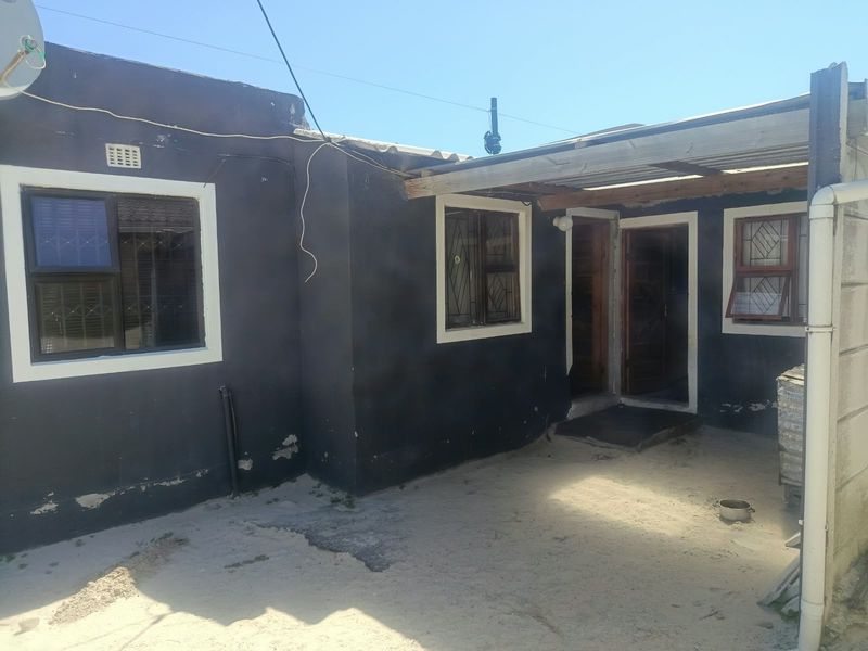 Rear exterior view of the property featuring a covered patio area, dark painted walls with security-fitted windows, and an unpaved sandy courtyard.