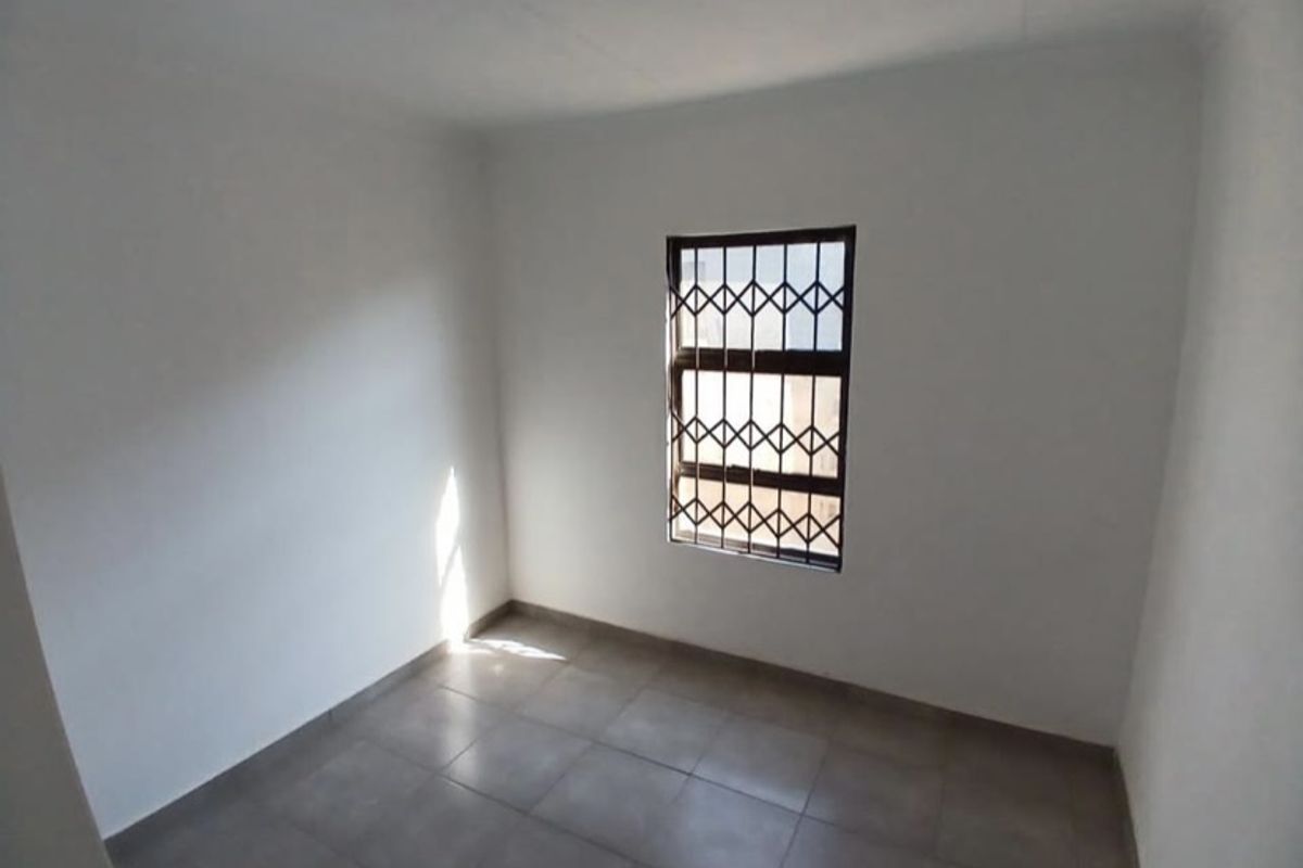 A clean, empty bedroom featuring neutral tiled flooring and a window fitted with a security gate.
