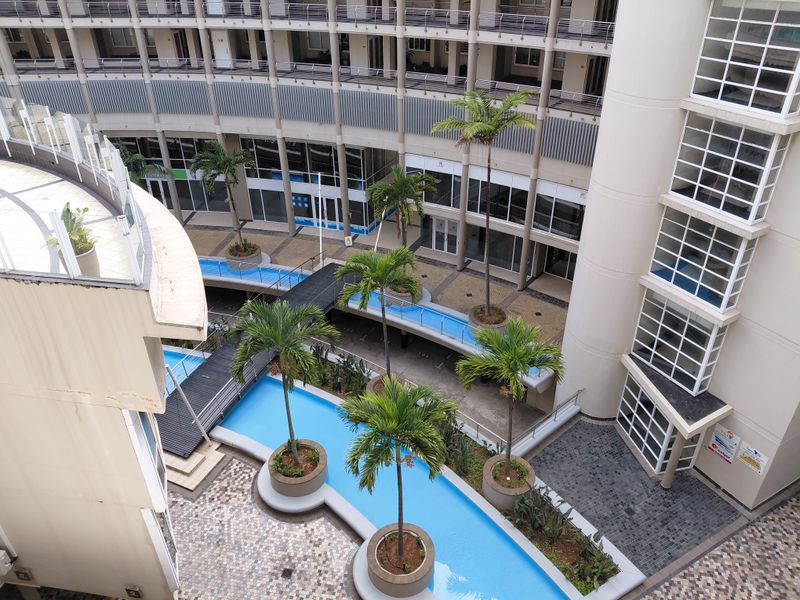 An elevated view of a modern apartment complex courtyard featuring a central water feature, landscaped palm trees, and paved pedestrian walkways.