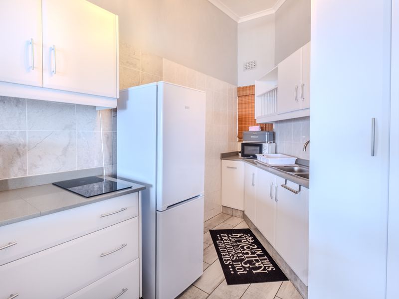 A compact, well-maintained kitchen featuring white cabinetry, stone countertops, an integrated electric hob, and neutral tiled finishes throughout.