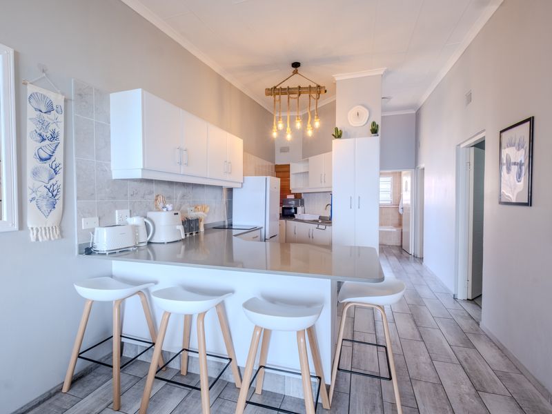 A modern, well-lit kitchen featuring white cabinetry, stone countertops, a tiled backsplash, and a breakfast bar with pendant lighting.