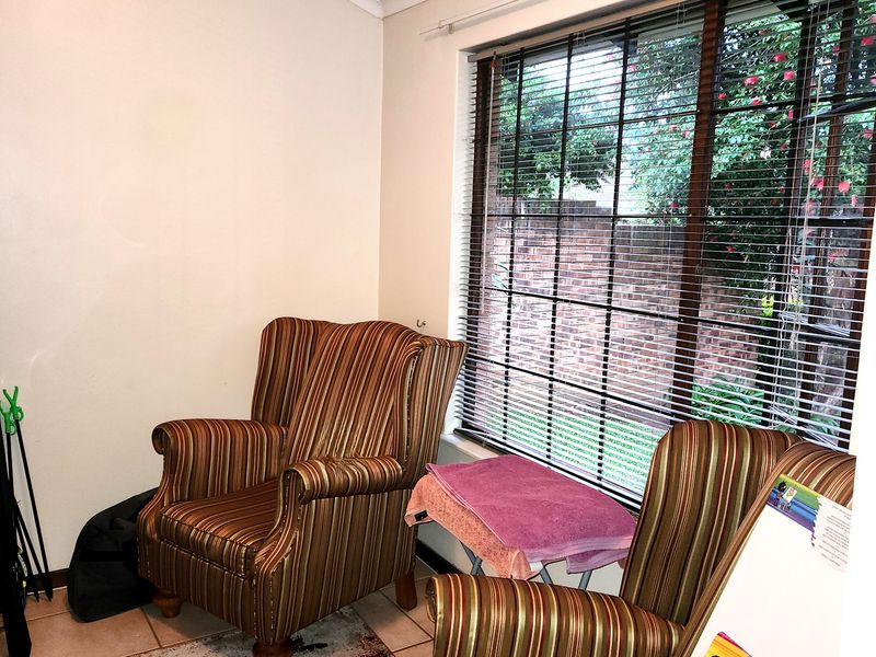 A living area featuring tiled flooring, coved ceilings, and large windows fitted with venetian blinds, offering a view of the garden.