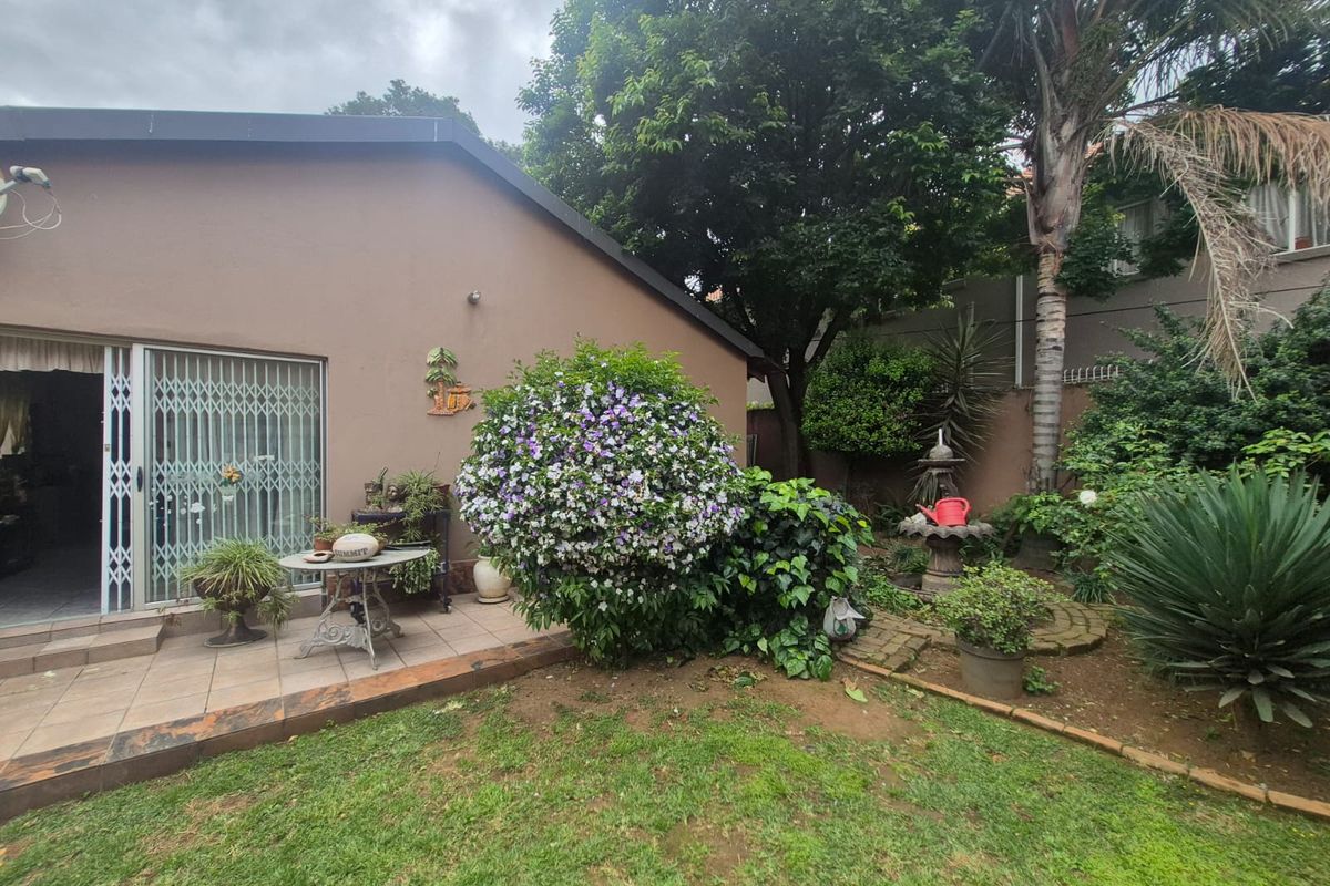 A rear view of the property featuring a tiled patio area, sliding glass doors with security gates, and a well-established landscaped garden with a fountain.