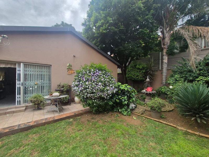 A rear view of the property featuring a tiled patio area, sliding glass doors with security gates, and a well-established landscaped garden with a fountain.