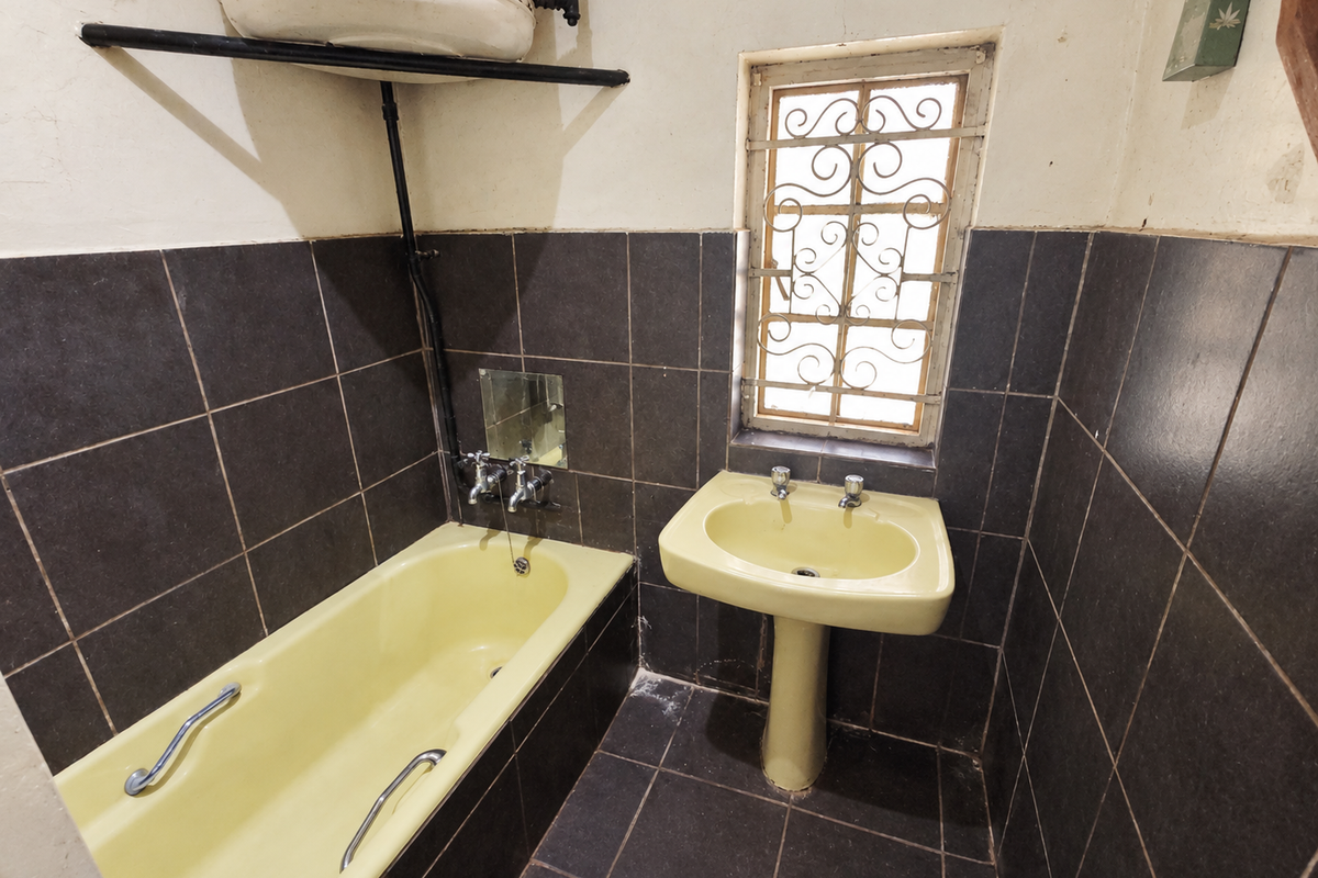 A dated bathroom featuring a yellow pedestal basin and bathtub, dark wall tiles, and an exposed wall-mounted geyser unit.