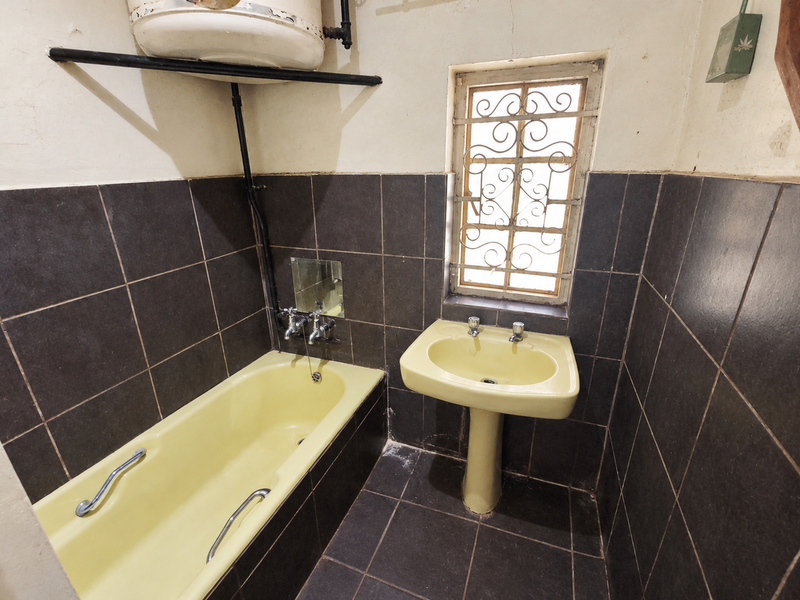 A dated bathroom featuring a yellow pedestal basin and bathtub, dark wall tiles, and an exposed wall-mounted geyser unit.