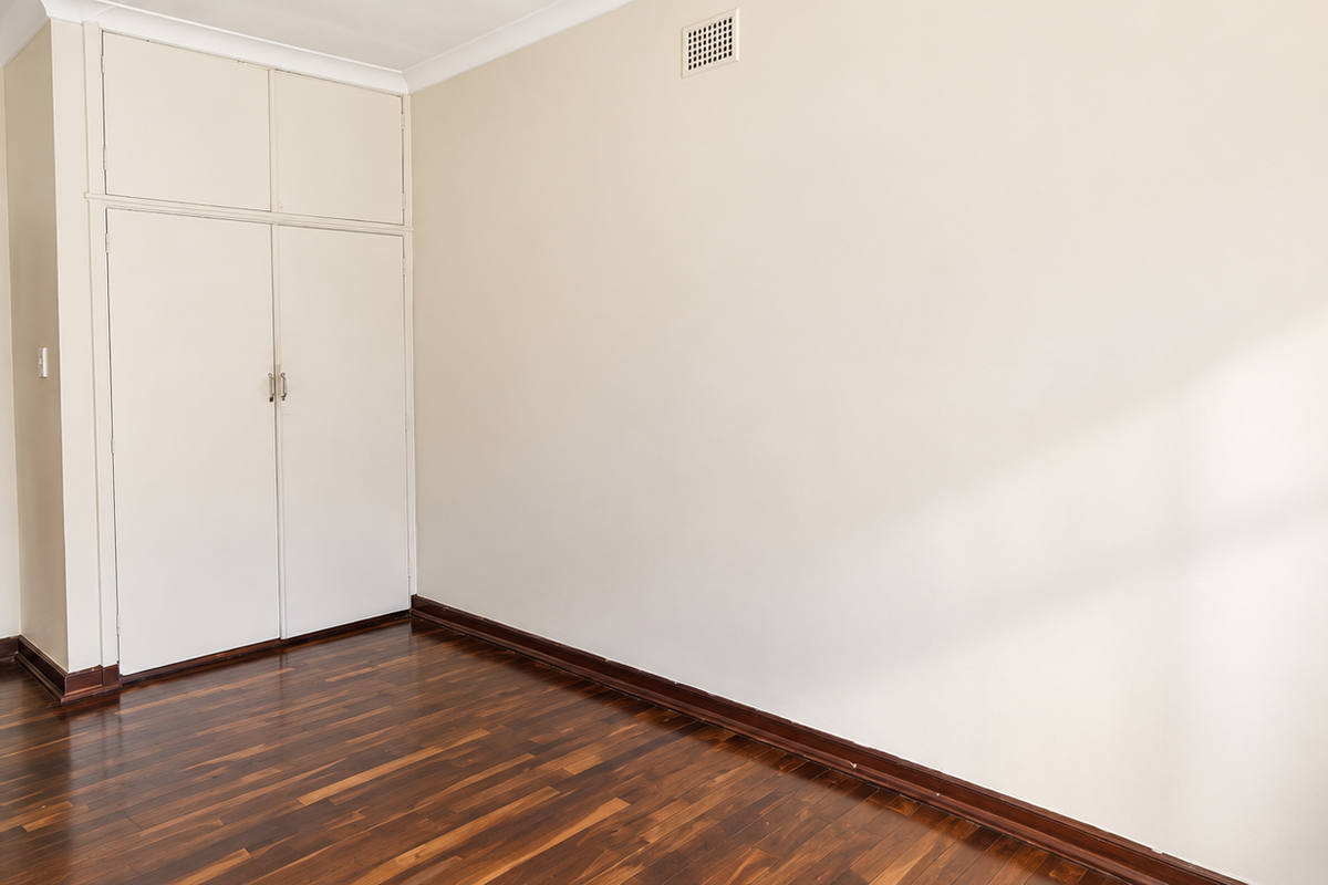 A bright, empty bedroom featuring polished timber flooring, built-in storage cabinetry, and neutral wall finishes.