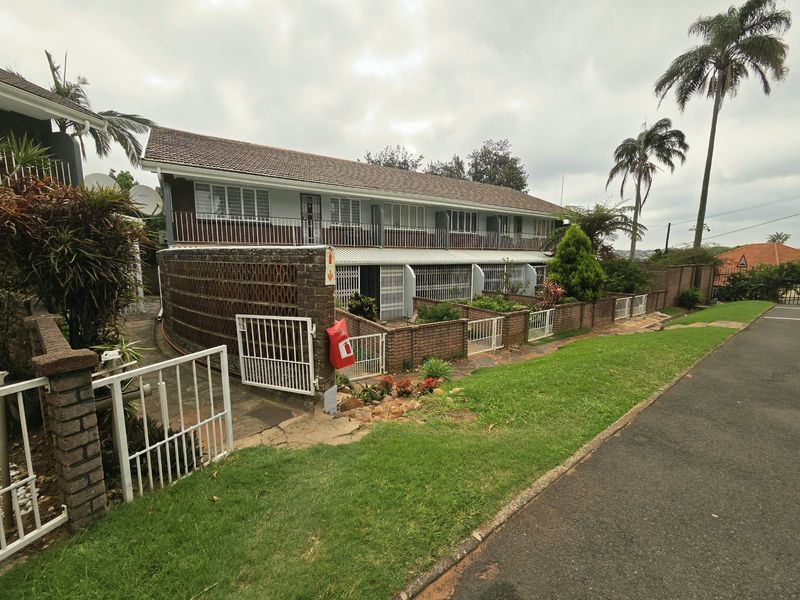 Front elevation of a multi-unit townhouse complex featuring brick boundary walls, security gates, and a tiled roof structure.