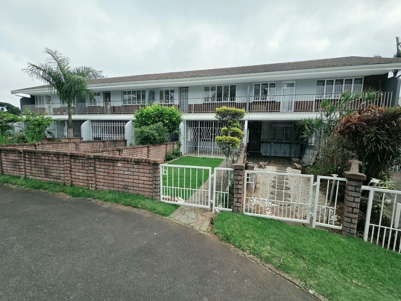 Front elevation of a townhouse complex featuring a brick boundary wall, security gates, and a shared upper-level balcony with metal railings.