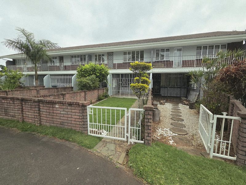 Front view of a two-story townhouse complex featuring a private garden, brick boundary walls, security gates, and an upper-level balcony.