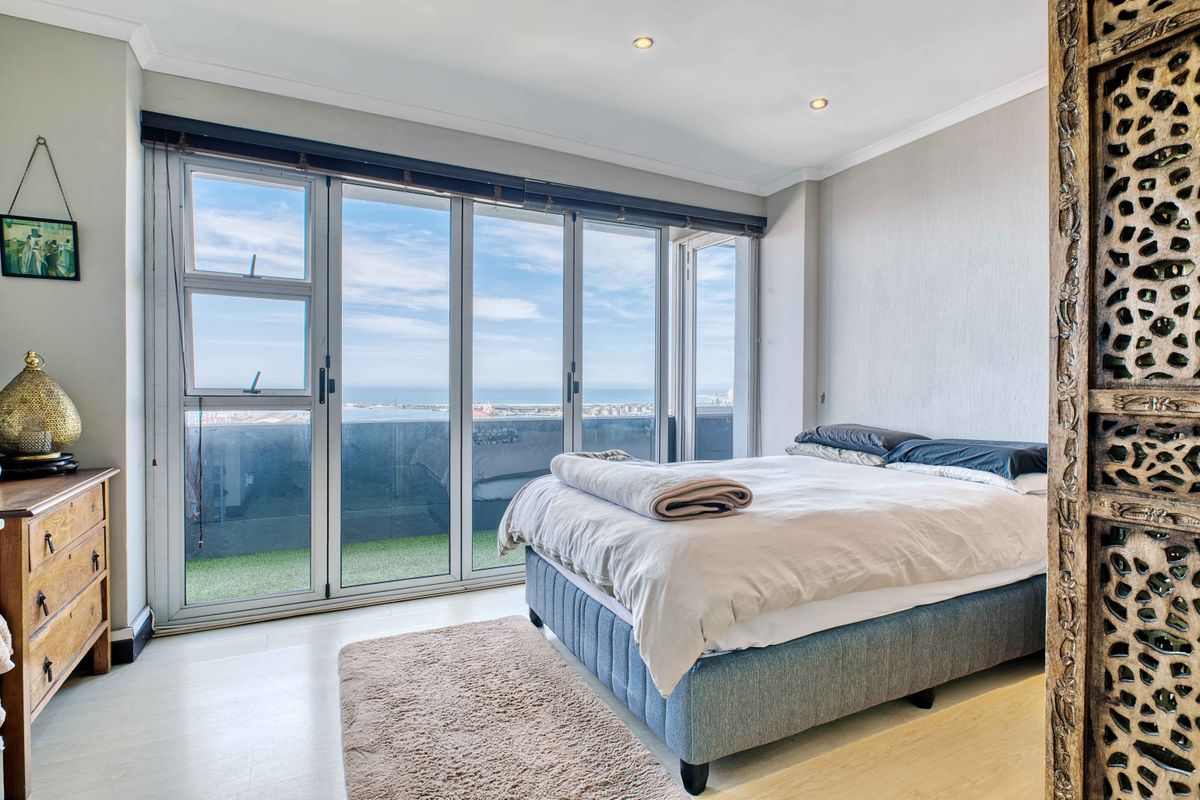 A bright bedroom featuring laminate flooring, recessed lighting, and large aluminium stacker doors opening onto a private balcony with expansive coastal views.