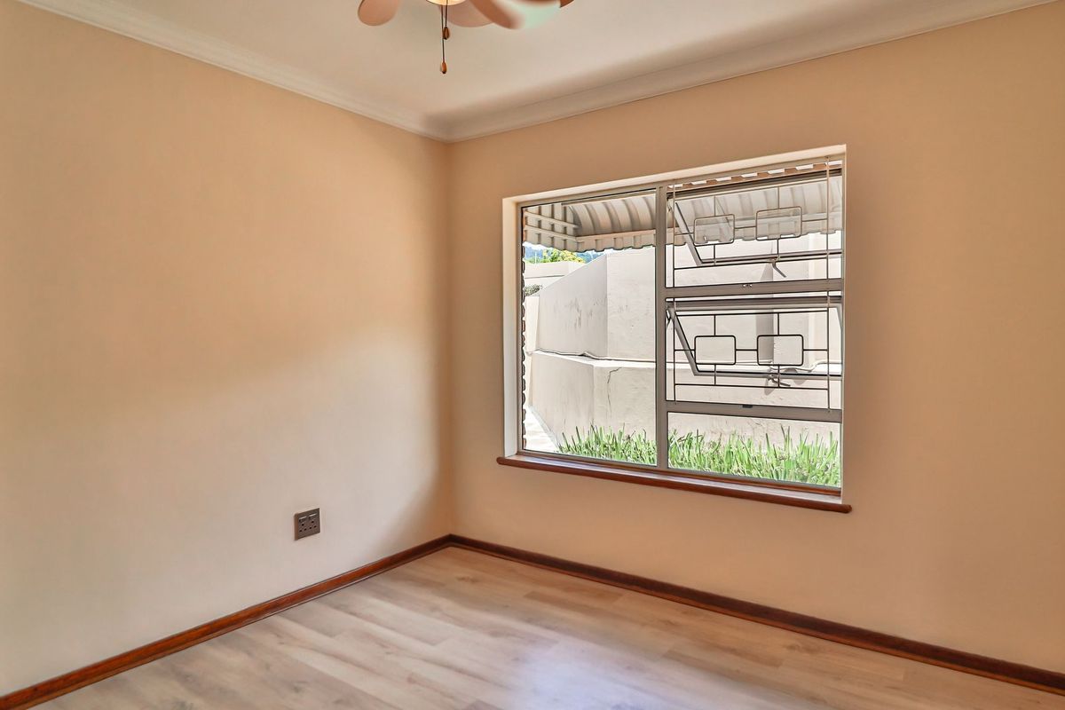 A bright, empty bedroom featuring laminate flooring, a ceiling fan, and an aluminium window with security bars, offering a clean and neutral living space.