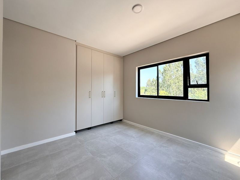 A bright, empty bedroom featuring neutral wall tones, modern grey floor tiles, and built-in cupboards with a view of the surrounding greenery.