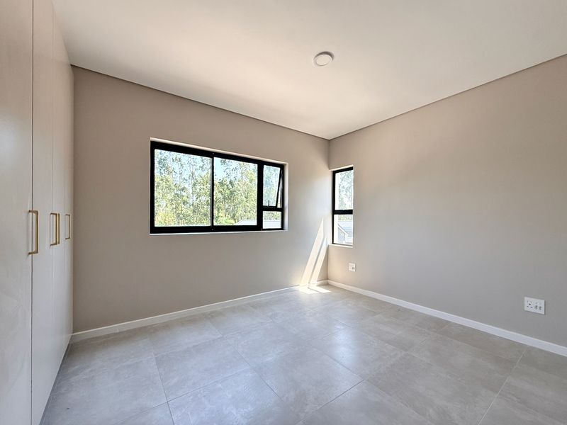 A bright, modern, and empty bedroom featuring neutral tiled flooring, built-in cupboards, and large aluminium-framed windows allowing for ample natural light.