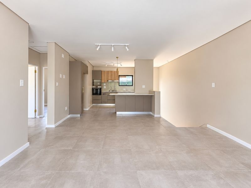 A spacious, modern open-plan living and kitchen area featuring neutral tiled flooring, contemporary cabinetry, and integrated kitchen appliances in a clean, vacant setting.