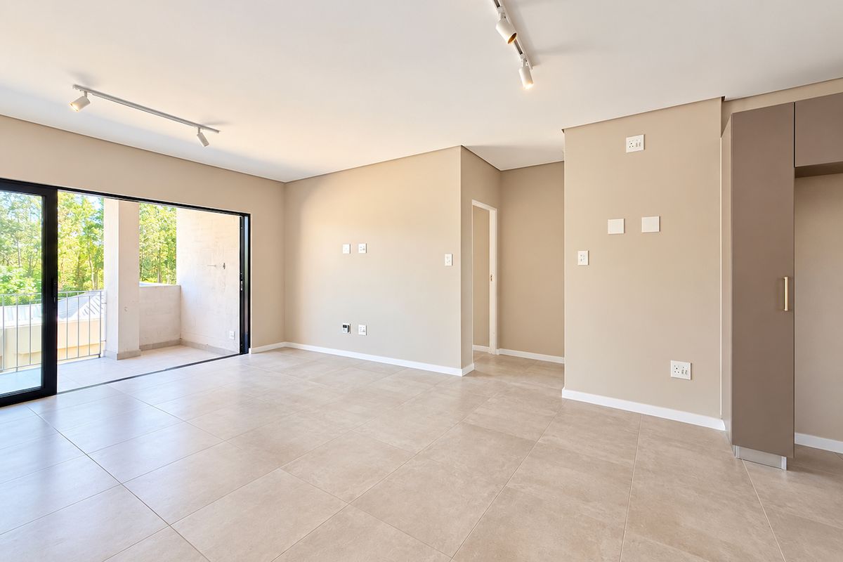 A bright, modern, and empty living area featuring neutral tiled flooring, track lighting, and a large sliding glass door leading to a private balcony.