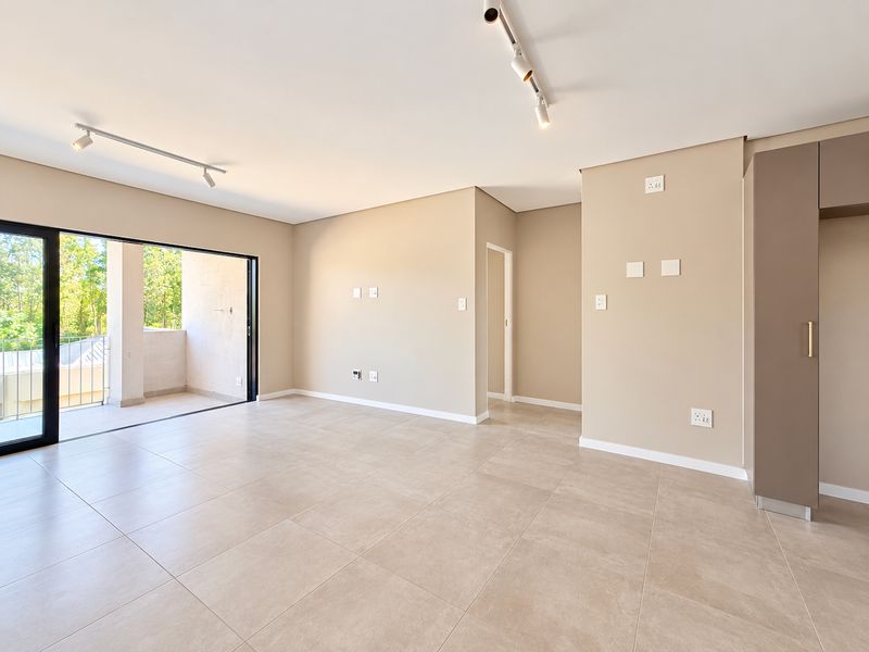 A bright, modern, and empty living area featuring neutral tiled flooring, track lighting, and a large sliding glass door leading to a private balcony.