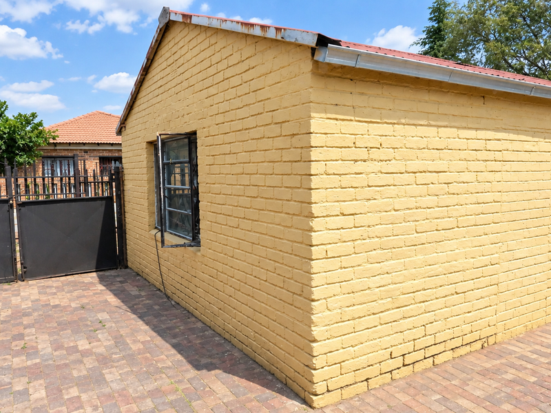 Exterior view of a painted brick outbuilding featuring a metal roof, security gate, and paved driveway area.