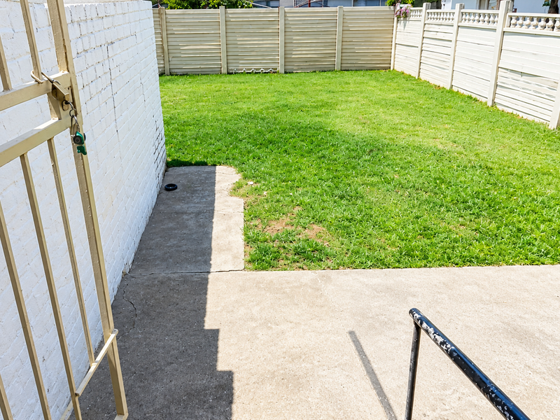 A private rear garden featuring a concrete patio area, a small lawn, and secure perimeter fencing with a painted brick wall.