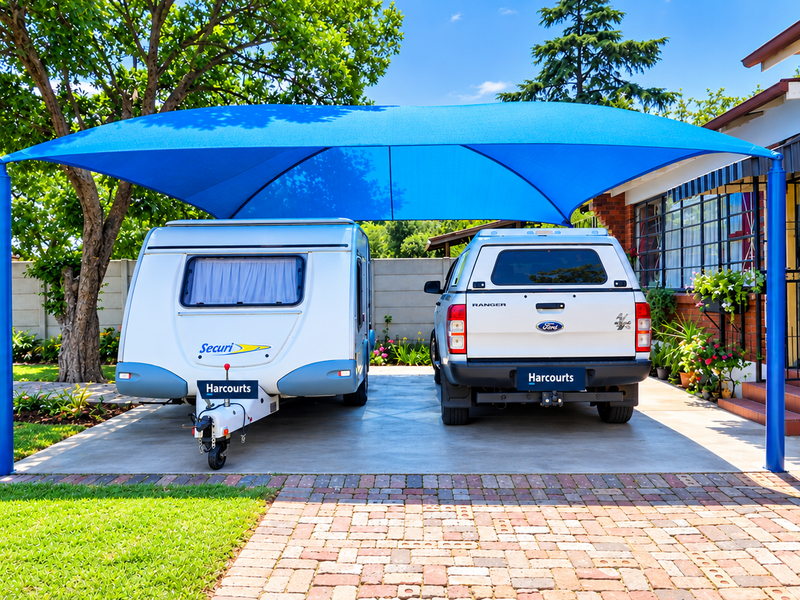 Front exterior view featuring a large blue shade sail covering a concrete driveway, adjacent to a brick house with security features.