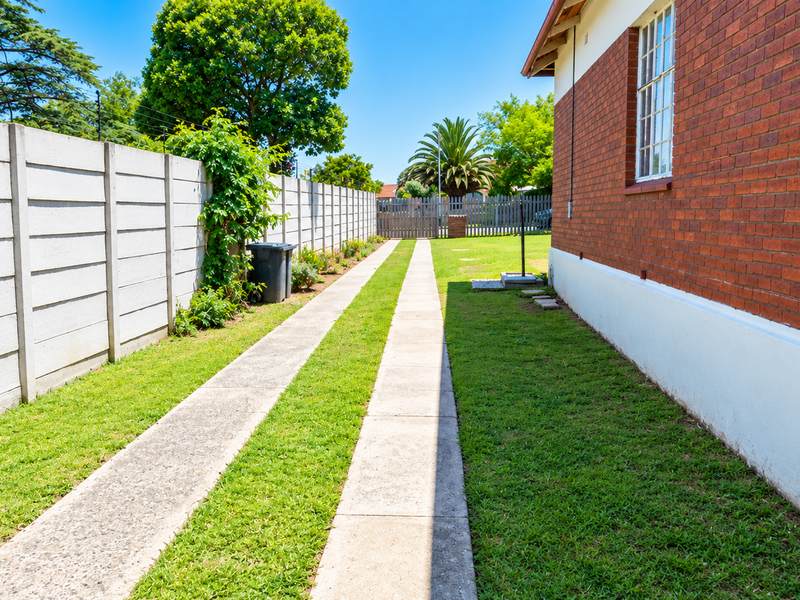 A side view of the property featuring a brick exterior wall, concrete driveway strips, and a precast concrete boundary wall enclosing the garden area.
