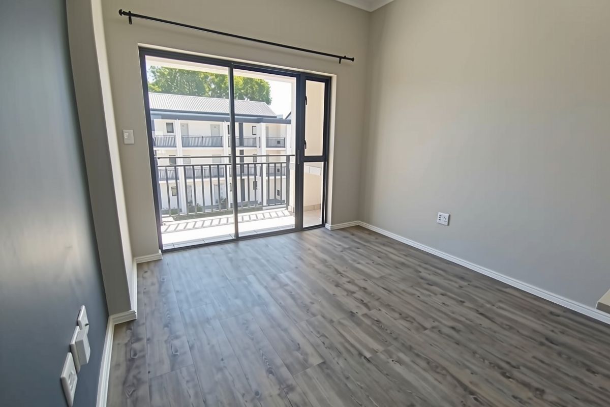 A bright, empty bedroom featuring modern laminate flooring, neutral walls, and a sliding glass door providing access to a private balcony.
