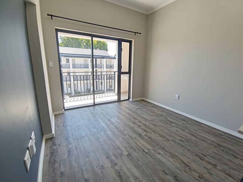 A bright, empty bedroom featuring modern laminate flooring, neutral walls, and a sliding glass door providing access to a private balcony.