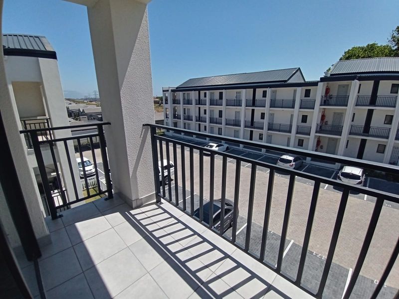 A clean, modern balcony with tiled flooring and a metal balustrade, overlooking the apartment complex parking area.