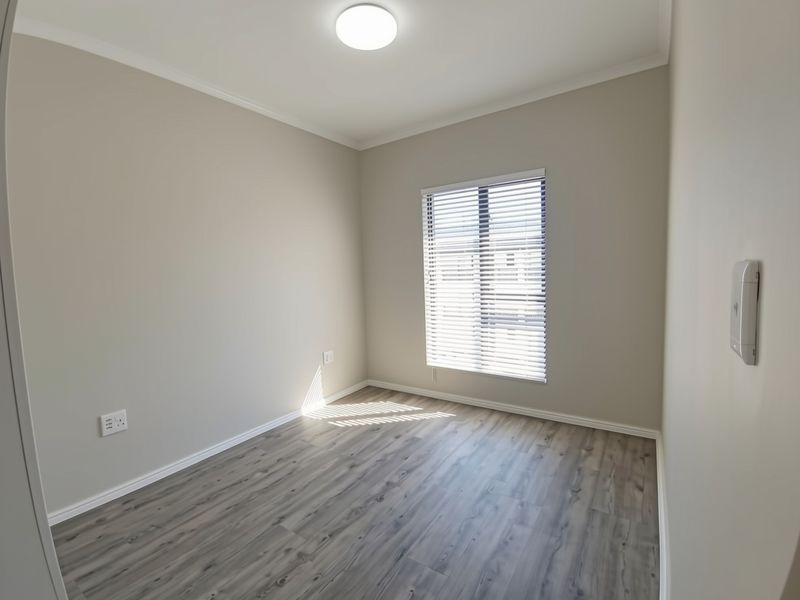 A bright, empty bedroom featuring modern laminate flooring, neutral wall tones, and a window fitted with venetian blinds.