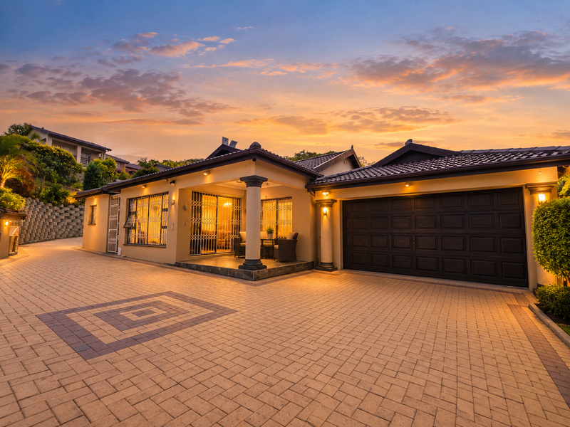 A well-maintained front exterior featuring a spacious paved driveway, double garage, and a welcoming covered patio under a tiled roof at sunset.