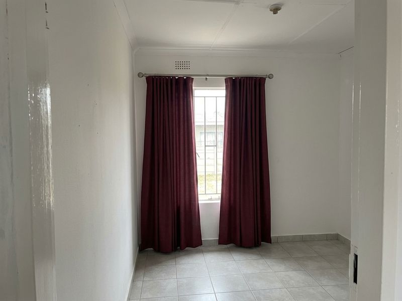 A vacant bedroom featuring neutral tiled flooring, white walls, and a window fitted with security bars and curtain rods.