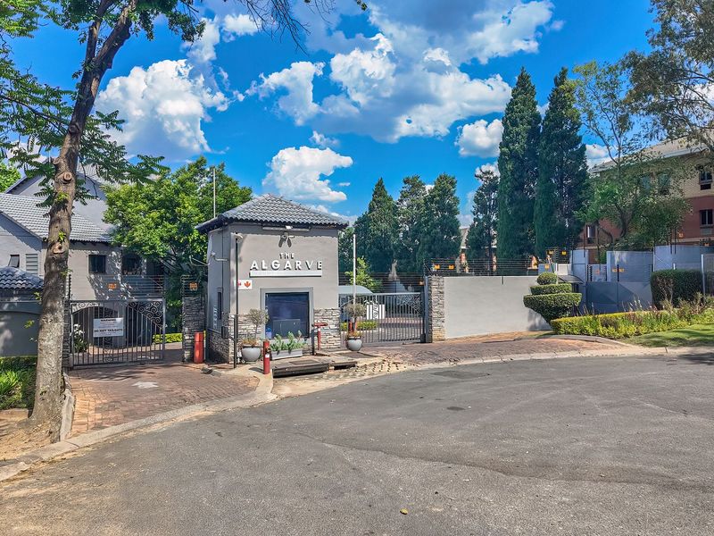 Front entrance view of The Algarve apartment complex featuring a secure guard house, gated access, and well-maintained perimeter fencing.