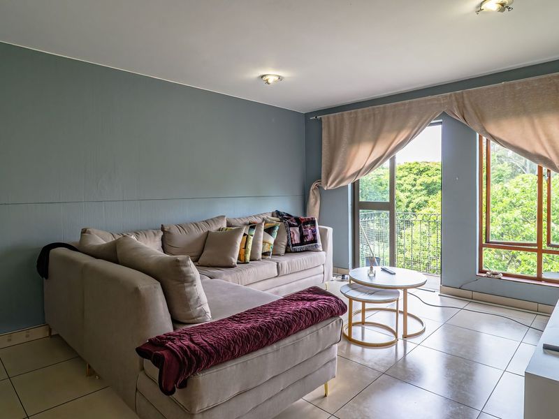 A living room featuring tiled flooring, large windows, and a sliding door providing access to a balcony with green views.