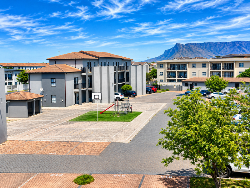 An elevated view of a townhouse complex featuring paved parking areas, a communal playground, and multi-story residential buildings with mountain views.