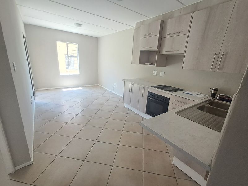 Open plan kitchen and living area featuring light wood-look cabinetry, built-in oven and hob, double sink, and neutral tiled flooring throughout.
