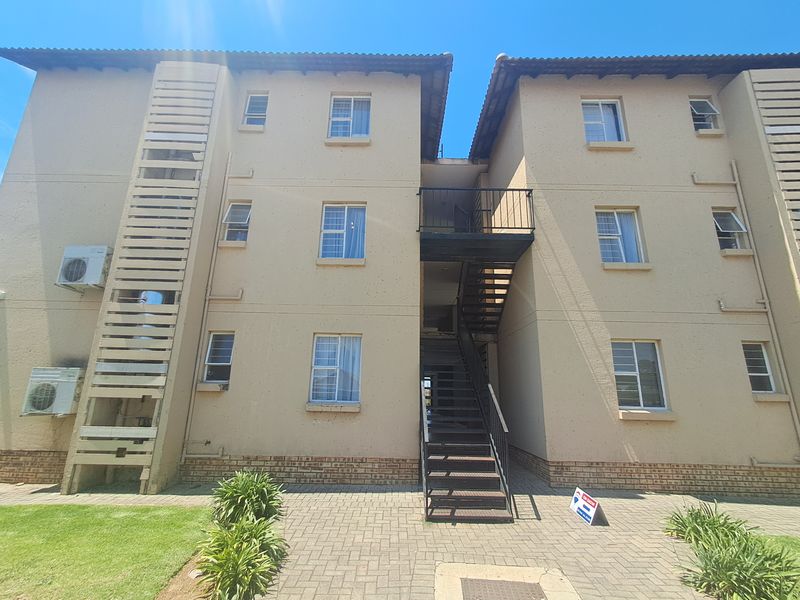 Exterior view of a multi-story apartment block featuring a central metal staircase, paved ground area, and external air conditioning units.