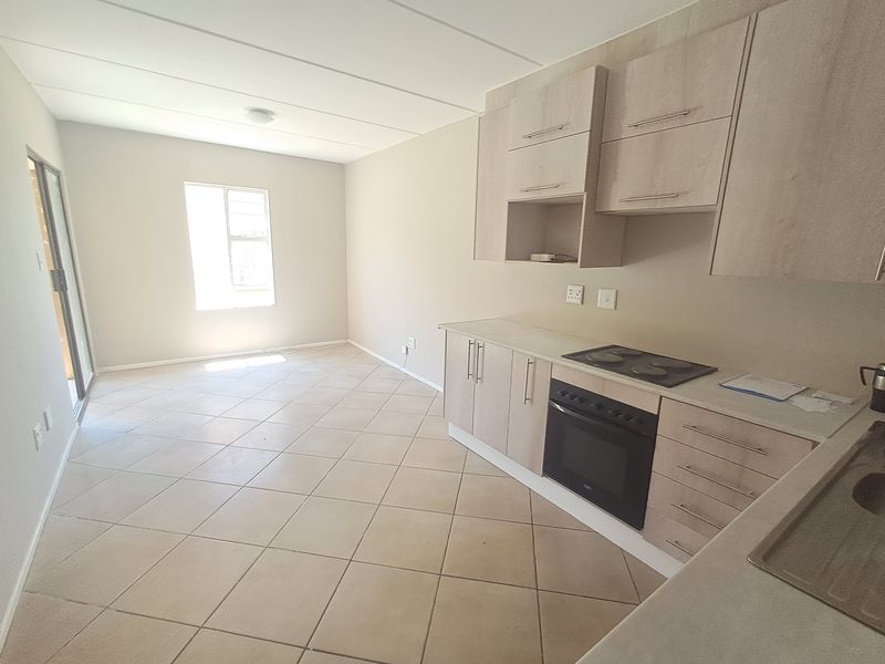 An open-plan living and kitchen area featuring light wood-finish cabinetry, built-in oven and hob, tiled flooring, and access to an outdoor space via sliding doors.