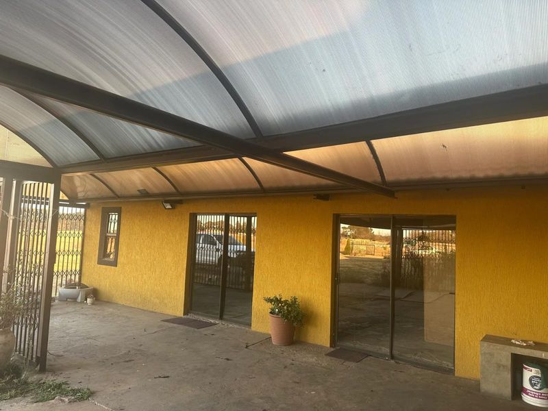 Covered patio area featuring a curved polycarbonate roof, concrete flooring, and sliding glass doors with security gates, providing access to the main dwelling.