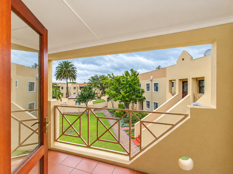 A covered balcony with tiled flooring and a metal balustrade, offering a pleasant view over the apartment complex courtyard and landscaped gardens.