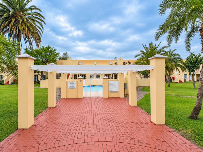 A well-maintained communal pool area featuring a paved walkway, pergola structure, and landscaped garden surroundings within the apartment complex.