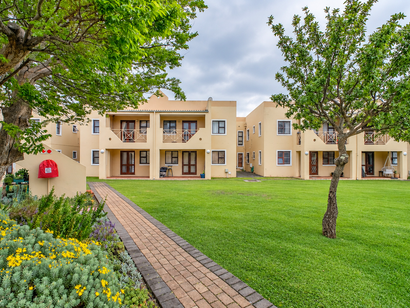 A well-maintained apartment complex exterior featuring a lush green lawn, paved walkway, and private balconies with sliding door access.