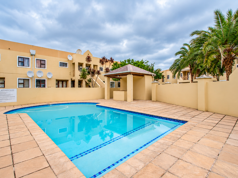 A well-maintained communal swimming pool area featuring a tiled deck, a covered gazebo, and surrounding perimeter walls within the apartment complex.