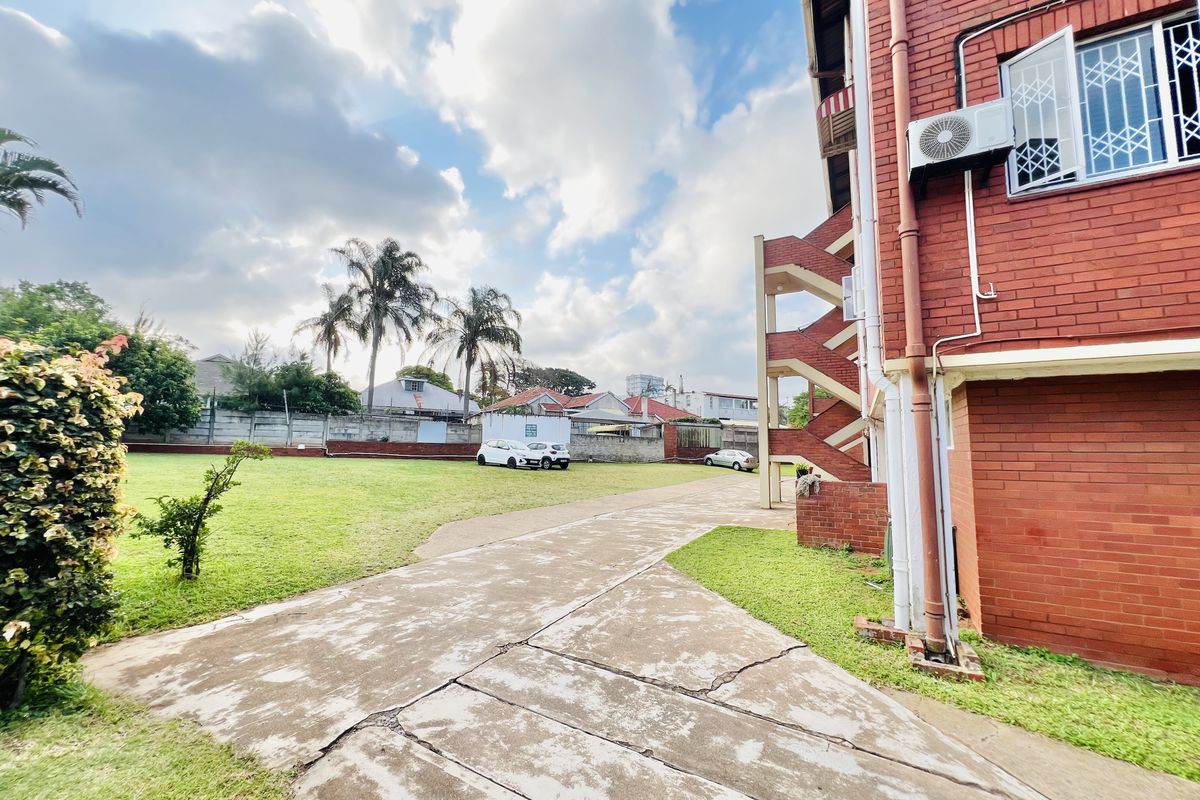 Rear view of a brick apartment building featuring an external staircase, security-barred windows, and a shared concrete driveway leading to a lawn area.