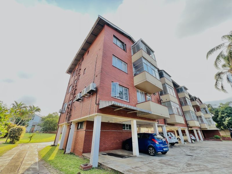 Exterior view of a multi-story red brick apartment building featuring covered ground-level parking bays and enclosed balconies on upper floors.