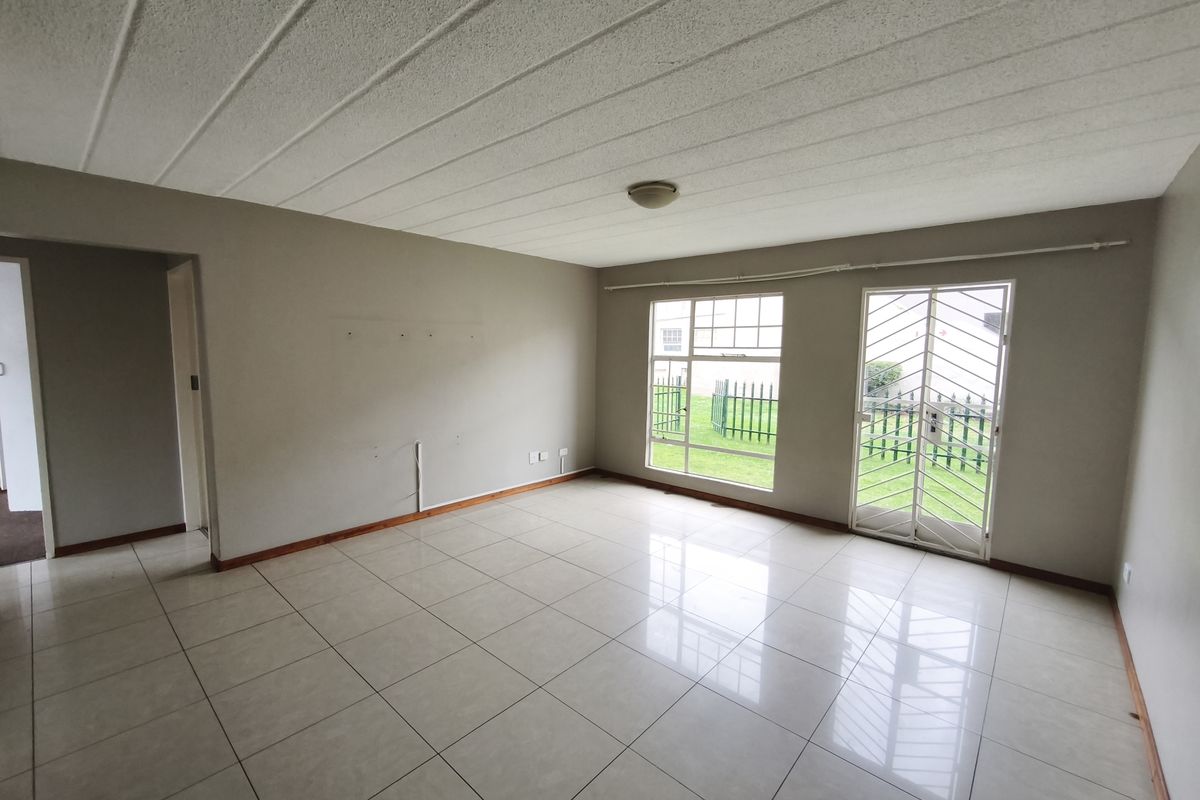 A spacious, empty living room featuring neutral tiled flooring, large windows for natural light, and a secure metal door leading to the exterior.