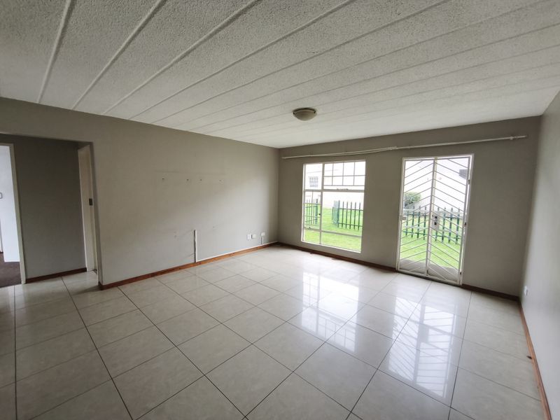 A spacious, empty living room featuring neutral tiled flooring, large windows for natural light, and a secure metal door leading to the exterior.
