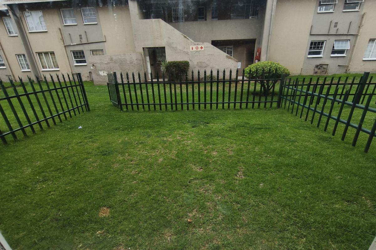 A view of the rear exterior of the apartment complex, featuring a private lawn area enclosed by metal security fencing.